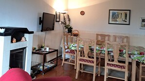 A cozy living room with a fireplace, a dining table with chairs, a television mounted on the wall, and a clock. - Vacation apartment for up to 8 people, 140 sqm, 2 WCs, directly on the L.Heide nature reserve (Bispingen)