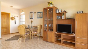 Una sala de estar con una mesa de comedor, sillas de mimbre, una vitrina de vidrio, un televisor y una estantería.