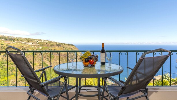 Una mesa de vidrio redonda con dos sillas de malla, una botella de vino y un frutero en un balcón con vistas al mar.