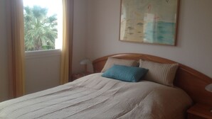 A neatly made bed with a blue pillow, a nightstand with a lamp, and a painting on the wall. - Family-friendly bungalow complex with a lot of green and a large pool (Miami Playa)