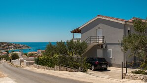 Una casa a due piani con un balcone, un'auto parcheggiata e vista sul mare e sulla cittadina costiera.