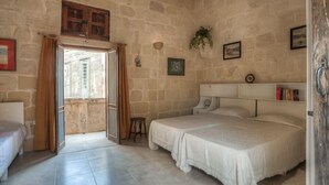 Un dormitorio con pared de piedra, cama, mesita de noche, ventana con cortinas y un taburete pequeño.
