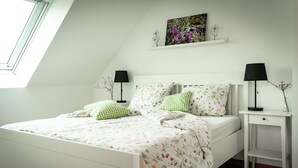 Une chambre dotée d’une lucarne, d’un lit avec une literie à motifs floraux, de deux tables de chevet avec des lampes et d’une étagère murale ornée d’œuvres d’art.