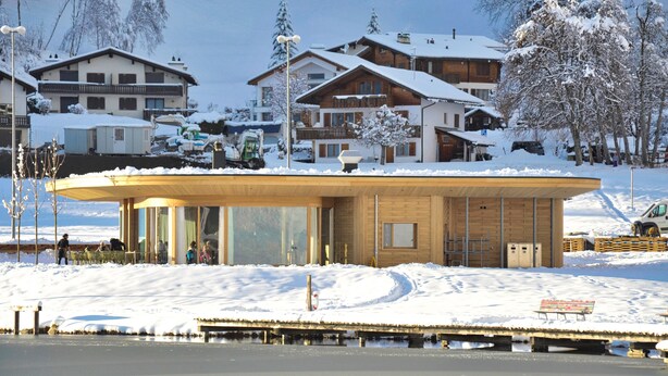 Un edificio moderno con grandes ventanales y fachada de madera, ubicado en un entorno nevado con casas al fondo.