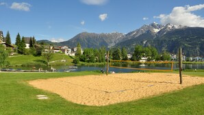 Una cancha de voleibol de arena con red, rodeada de césped verde y árboles, con un lago y montañas al fondo.