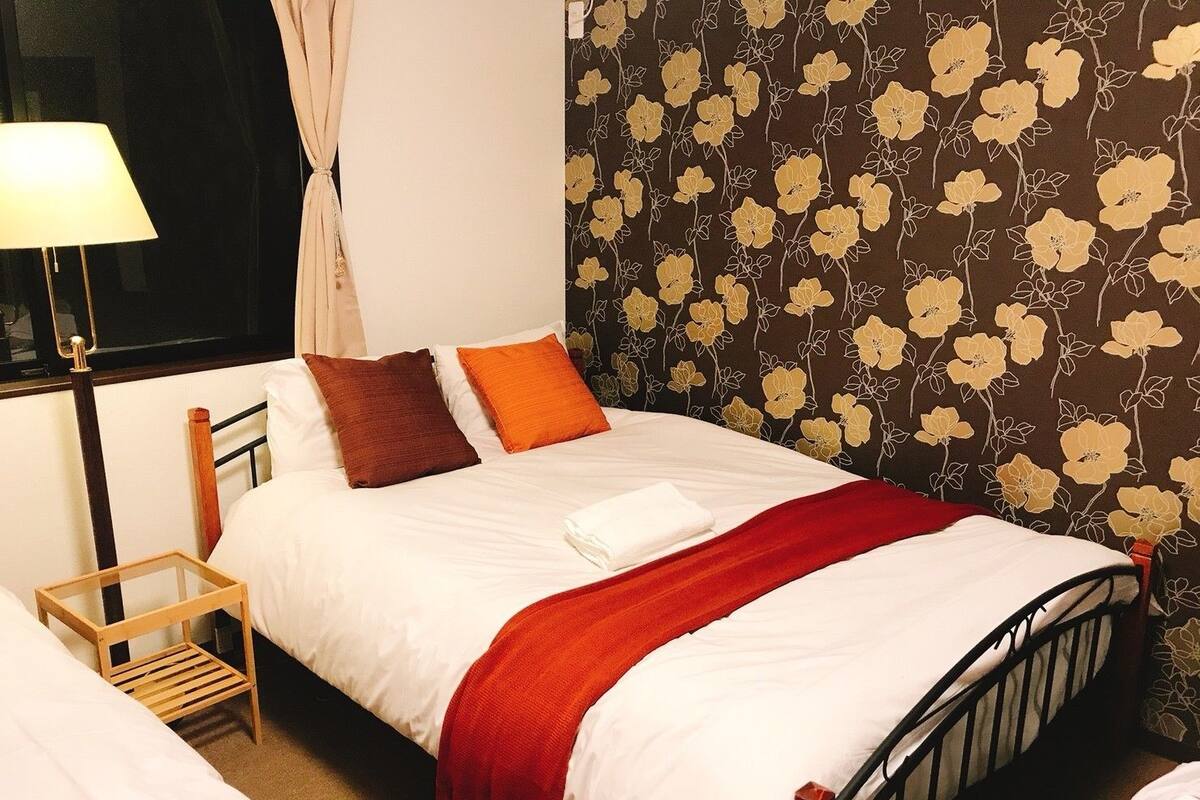 A neatly made bed with white bedding, two pillows, a red throw blanket, and a wooden nightstand with a lamp.