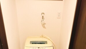 A compact, white, wall-mounted washing machine in a narrow hallway with a wooden door frame and a small window above it. - GH One More Heart at Toji 3 (Kyoto)