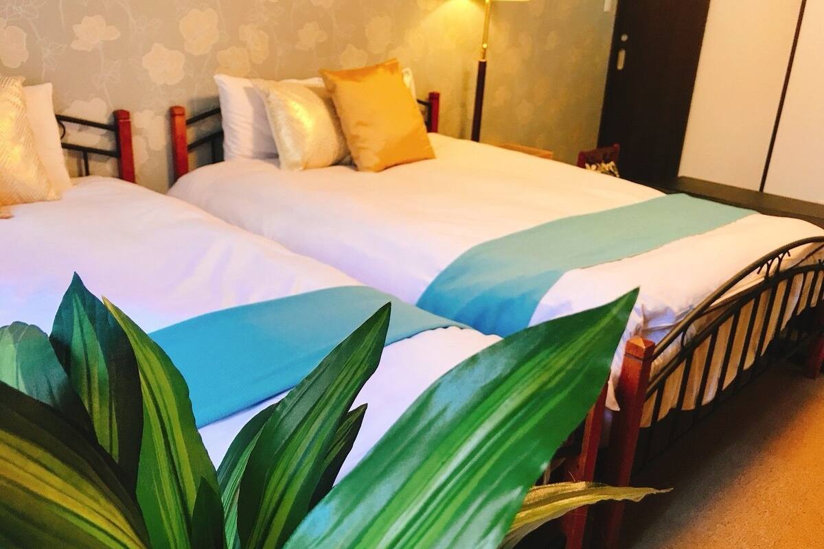A hotel room with two beds, a nightstand, a lamp, and a large plant in the foreground.