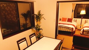 A hotel room with a dining table, chairs, a bed with red bedding, a nightstand, a lamp, and a potted plant. - GH One More Heart at Toji 3 (Kyoto)