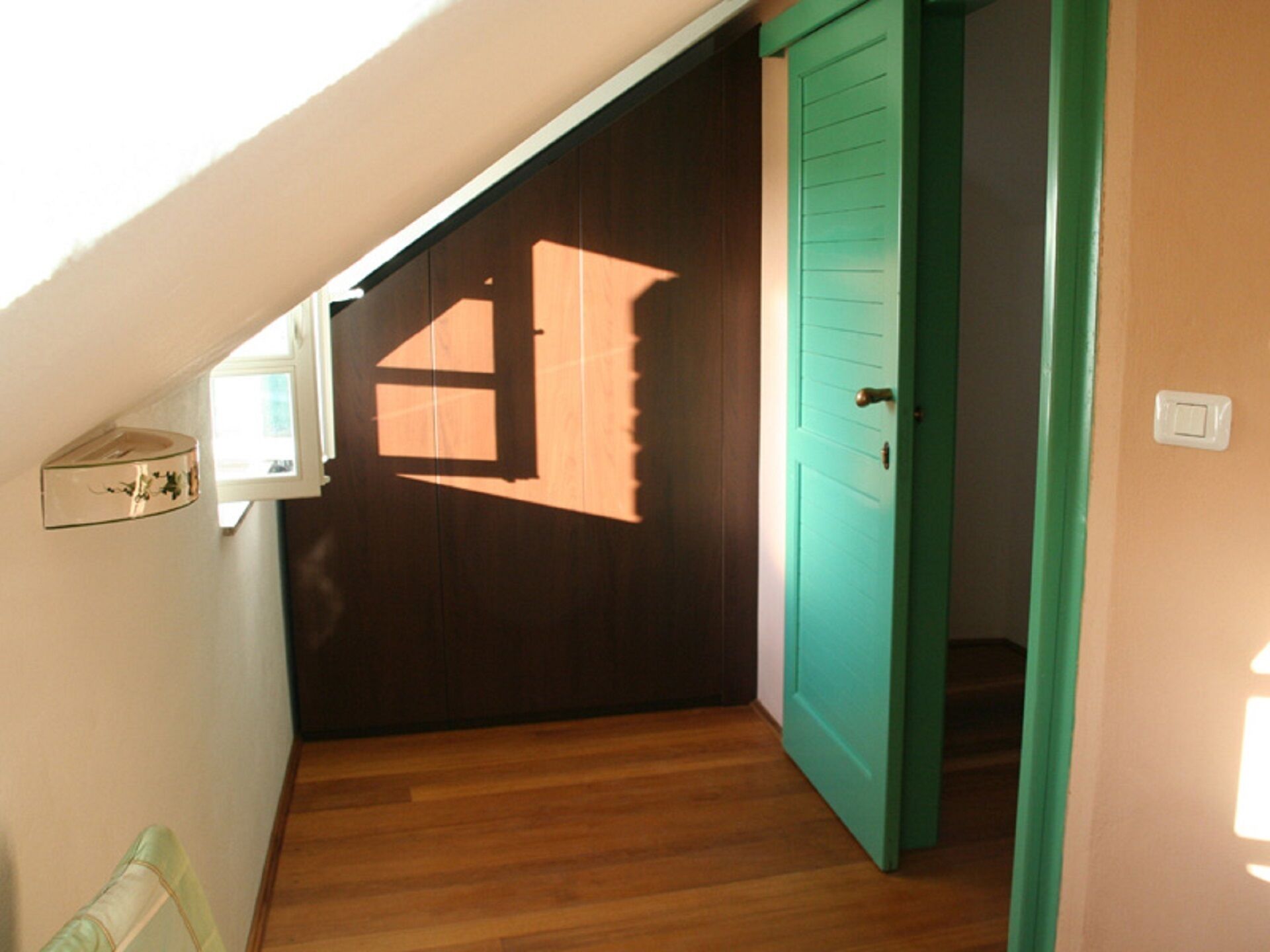 A narrow hallway with wooden flooring, a green door, and a window casting a shadow.