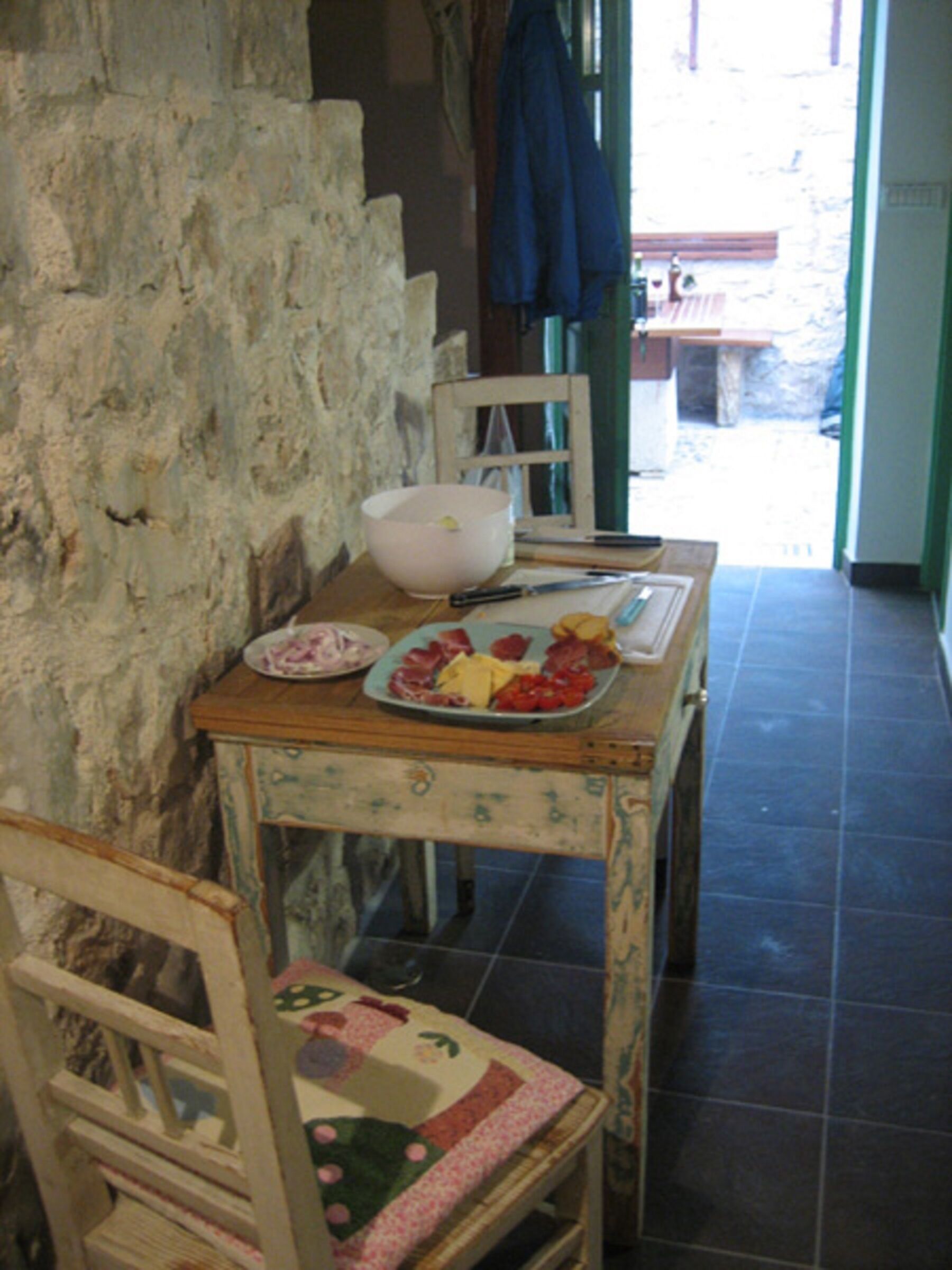 A rustic dining area with a stone wall, a wooden table with a plate of food, and a chair with a floral cushion.