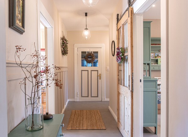 A hallway with a door, a hanging light fixture, a rug, and a vase with branches.