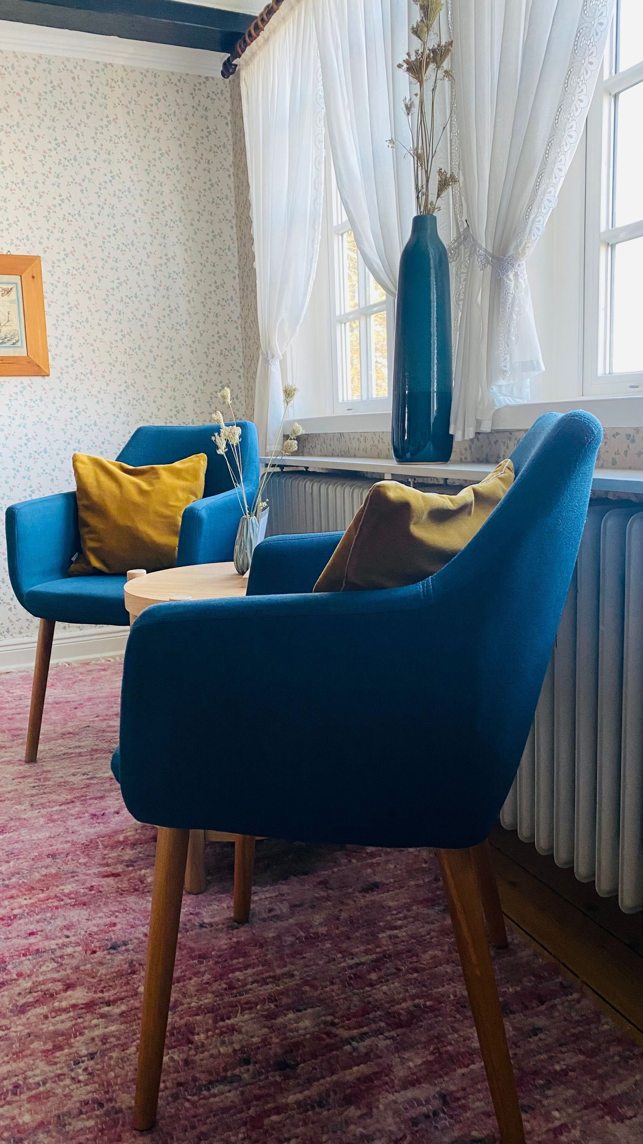 Ein Zimmer mit einem blauen Sessel, einem gelben Kissen, einer blauen Vase mit getrockneten Blumen und einem Fenster mit weißen Vorhängen.