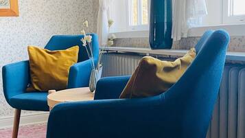 Ein Zimmer mit einem blauen Sessel, einem gelben Kissen, einer blauen Vase mit getrockneten Blumen und einem Fenster mit weißen Vorhängen.