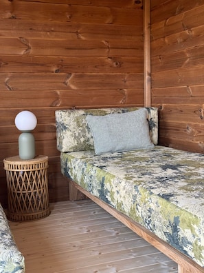Ein Holzblockhaus-Innenraum mit einem Bett, einem kleinen runden Weiden-Nachttisch und einer Wand mit Holzvertäfelung.