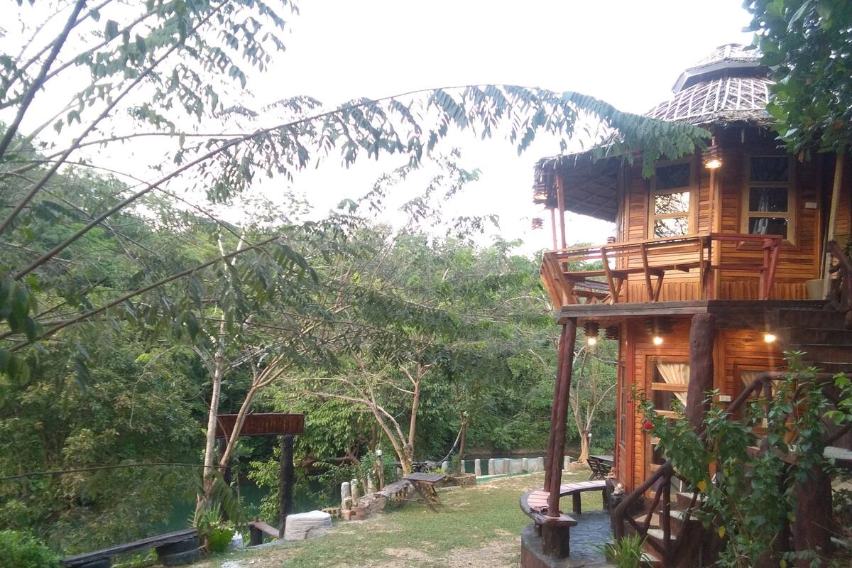 A wooden cabin with a balcony, surrounded by trees and a dirt path.
