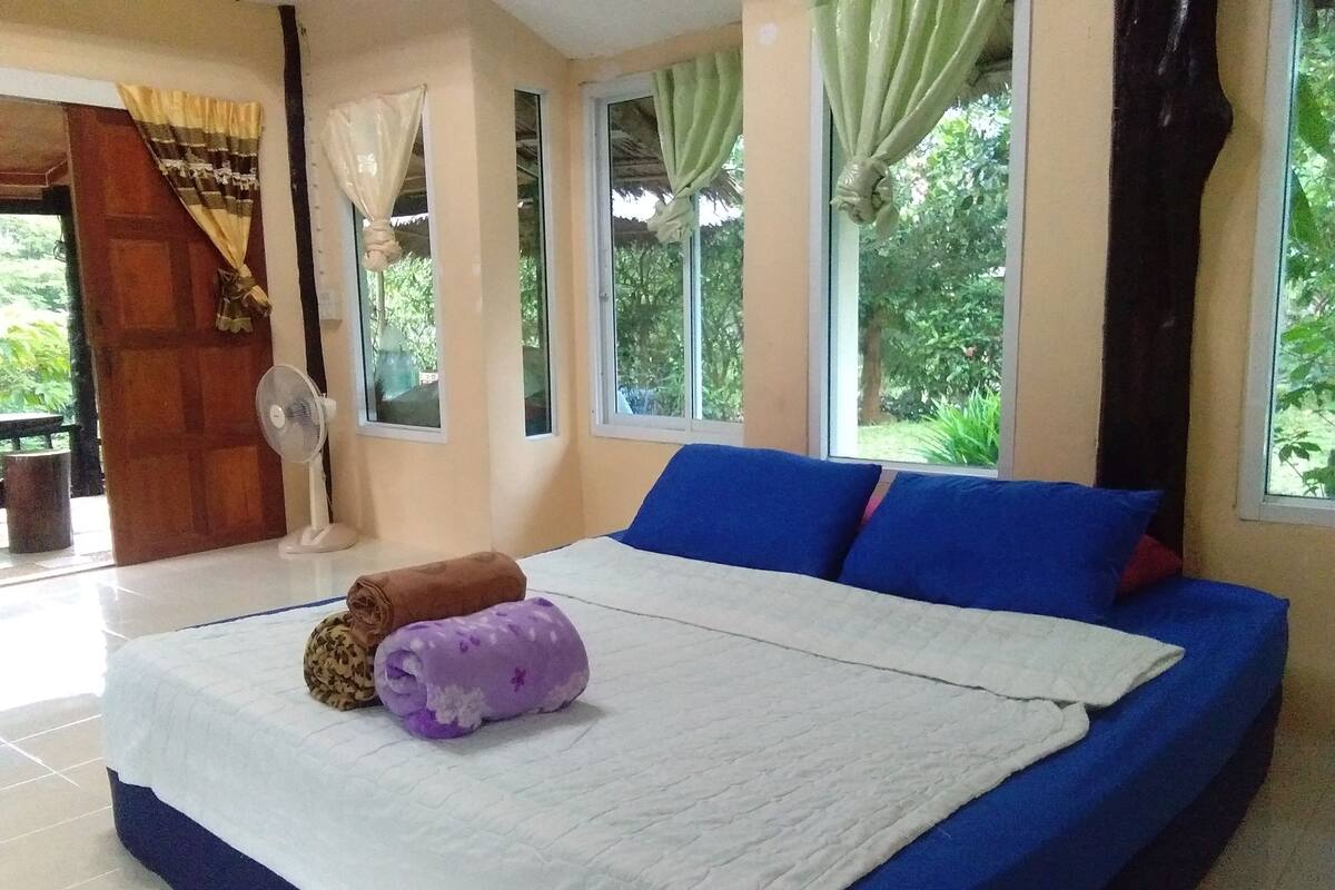 A bedroom with a bed, a fan, and a view of greenery outside.