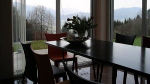 Un coin repas avec une table et des chaises, un vase rempli de fleurs et une vue sur les montagnes, visible à travers de grandes fenêtres.