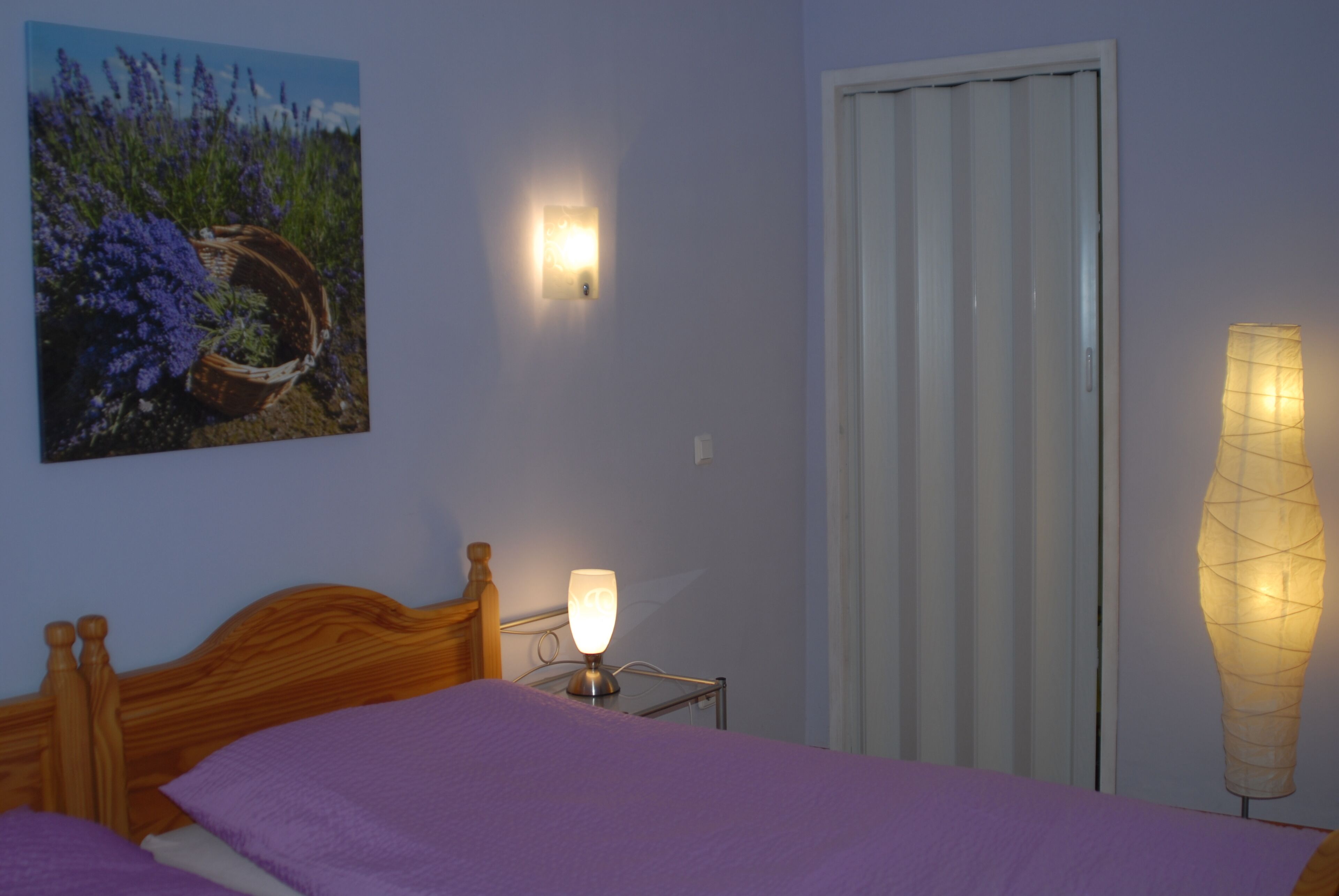 Ein Schlafzimmer mit einem Bett, einer Wandlampe, einem Lavendelbild und einem Fenster mit Jalousien.