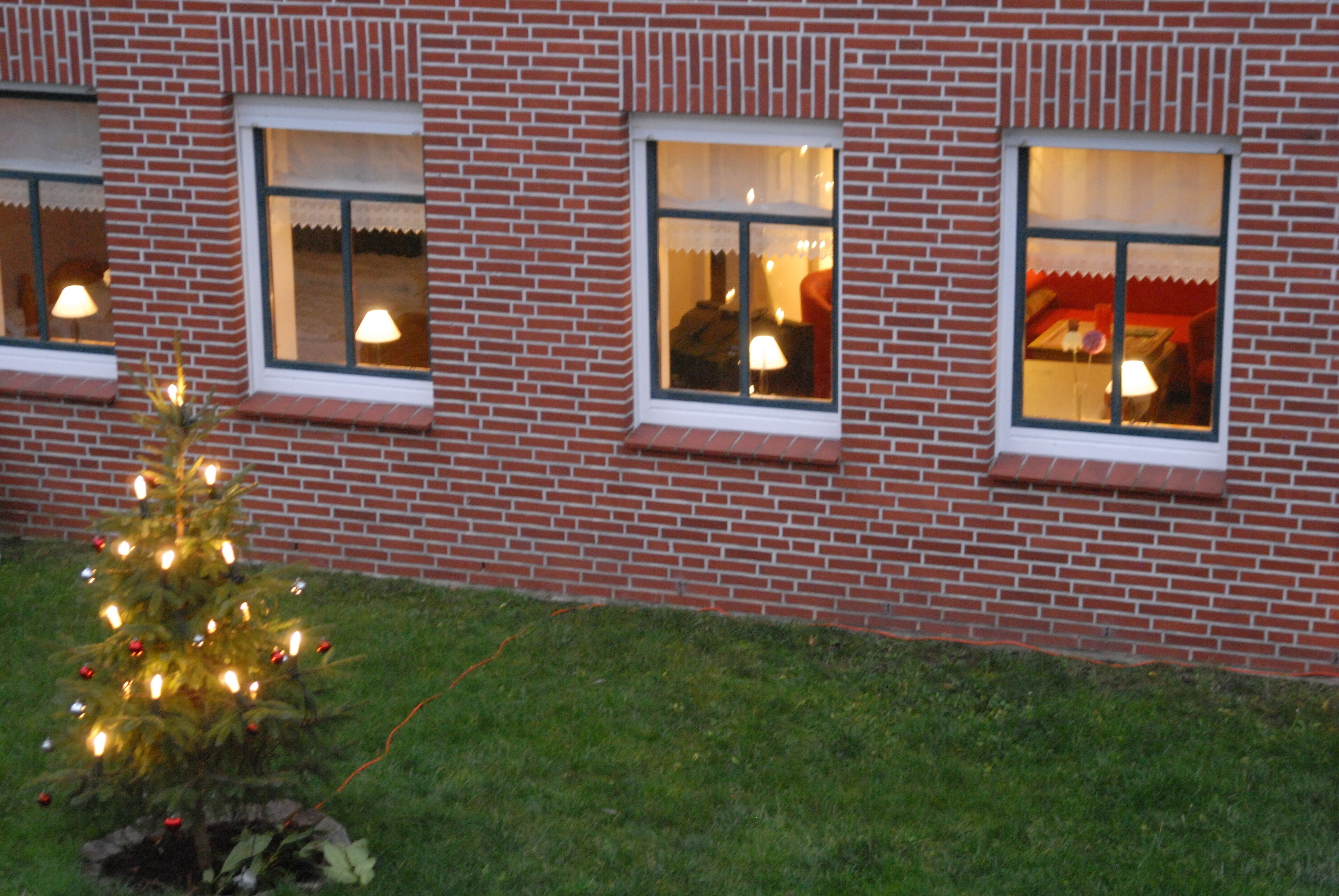 Ein Backsteinhaus mit drei Fenstern, jedes mit weißem Rahmen und Vorhängen. Vor dem Gebäude steht ein kleiner, beleuchteter Weihnachtsbaum.