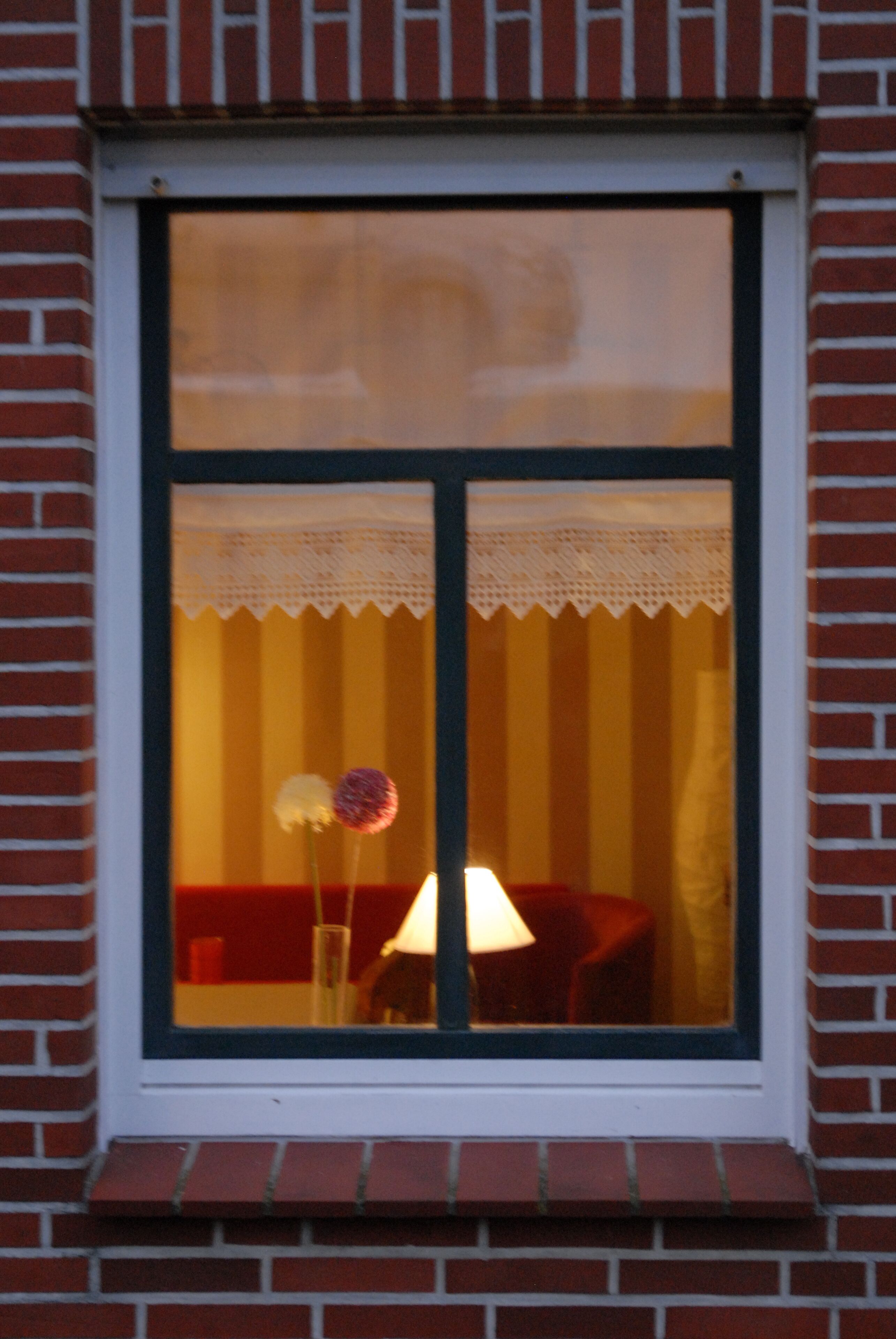 Une fenêtre donnant sur une pièce où l’on aperçoit une lampe, une chaise et une table.