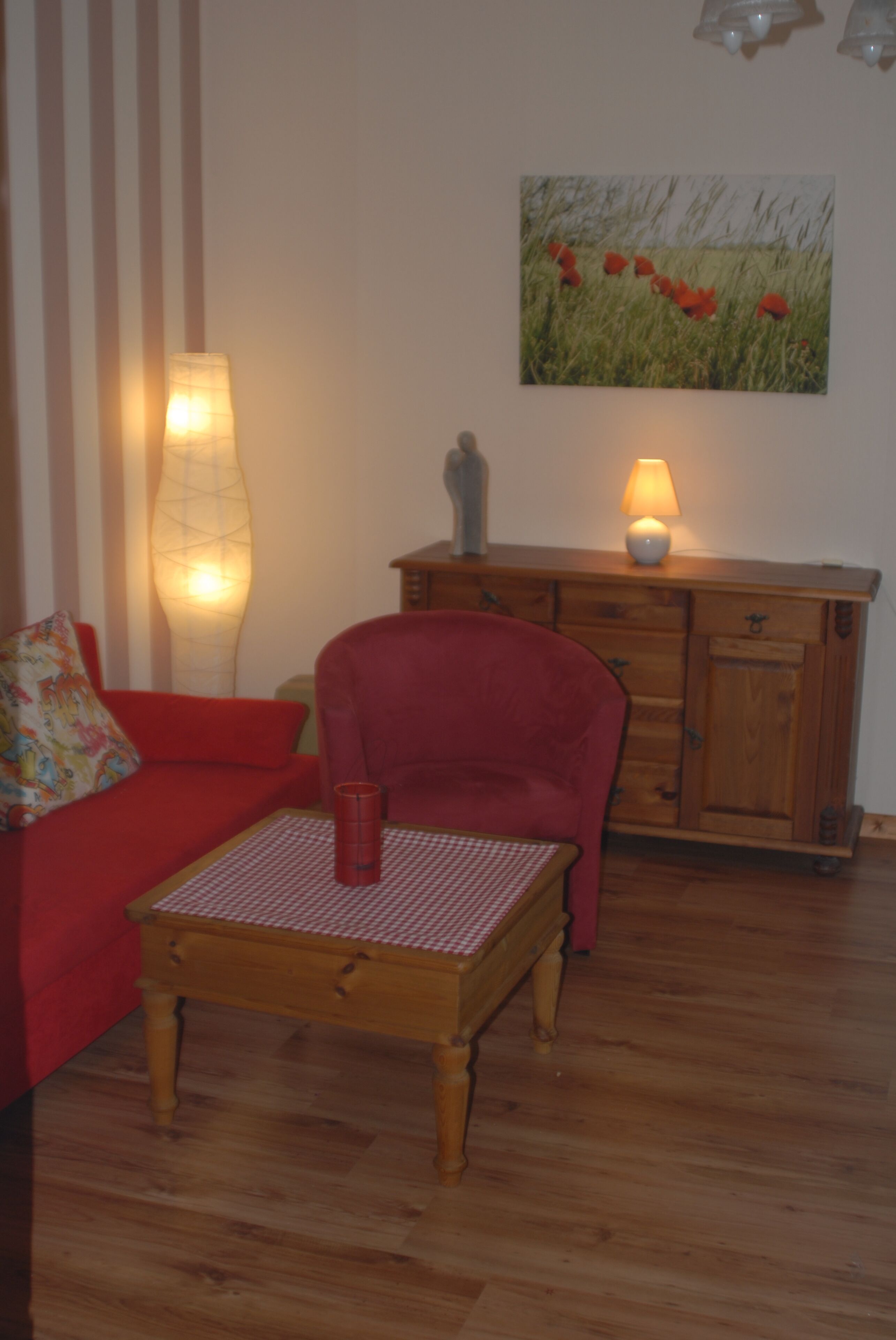 Ein Wohnzimmer mit einem roten Sofa, einem Holztisch, einer Lampe, einem Schrank und einem Gemälde an der Wand.