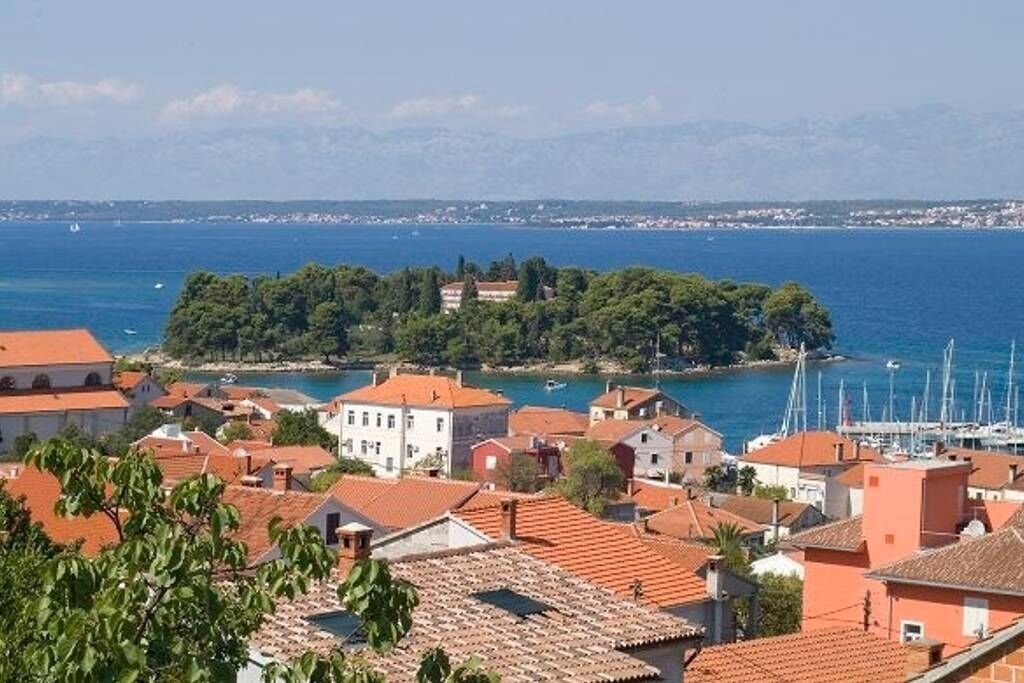 Une ville côtière aux bâtiments aux toits de tuiles rouges, un port de plaisance où sont amarrés des voiliers, et une vue dégagée sur la mer.