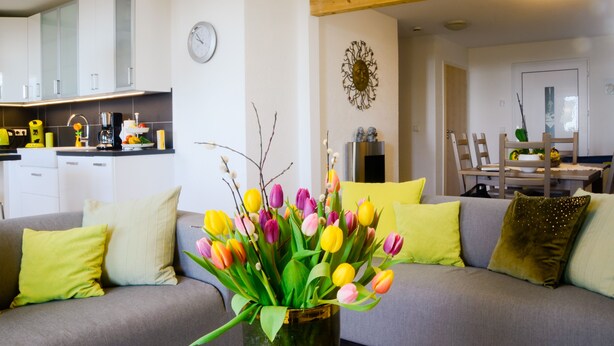 Una sala de estar moderna con un sofá gris, un florero con tulipanes de colores y un comedor con una mesa y sillas de madera.