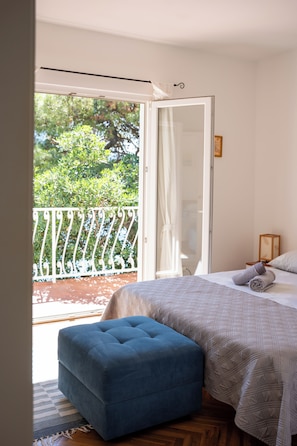 Une chambre Ă coucher avec un lit, un pouf bleu, une fenĂȘtre donnant sur un paysage verdoyant et une rambarde blanche.