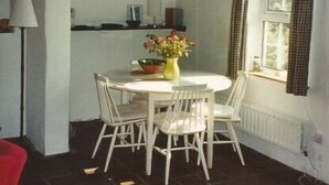 Un comedor con una mesa redonda y sillas, un jarrón con flores, una ventana con cortinas y un techo con vigas a la vista.