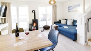 En moderne stue med en blå sofa, et træspisebord med vin og spillekort, en pejs og store vinduer med gardiner.