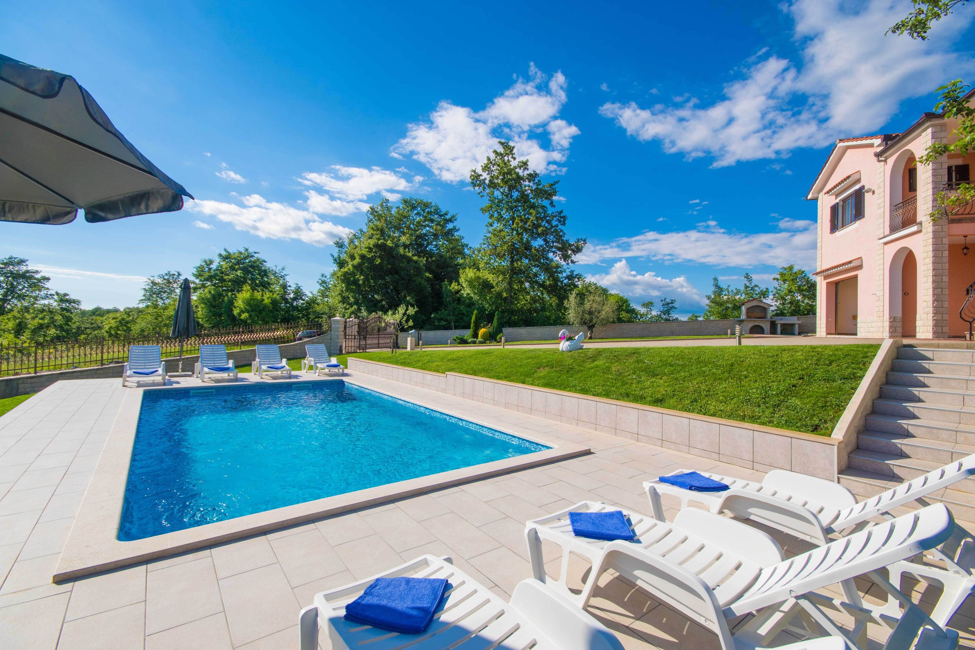 Ein rechteckiger Pool mit Liegestühlen und Sonnenschirmen, umgeben von einer gepflegten Rasenfläche und einem Gebäude mit Rundbogenfenstern.