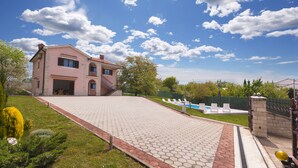 Uma casa de dois andares com um caminho de entrada pavimentado e uma piscina ao fundo.