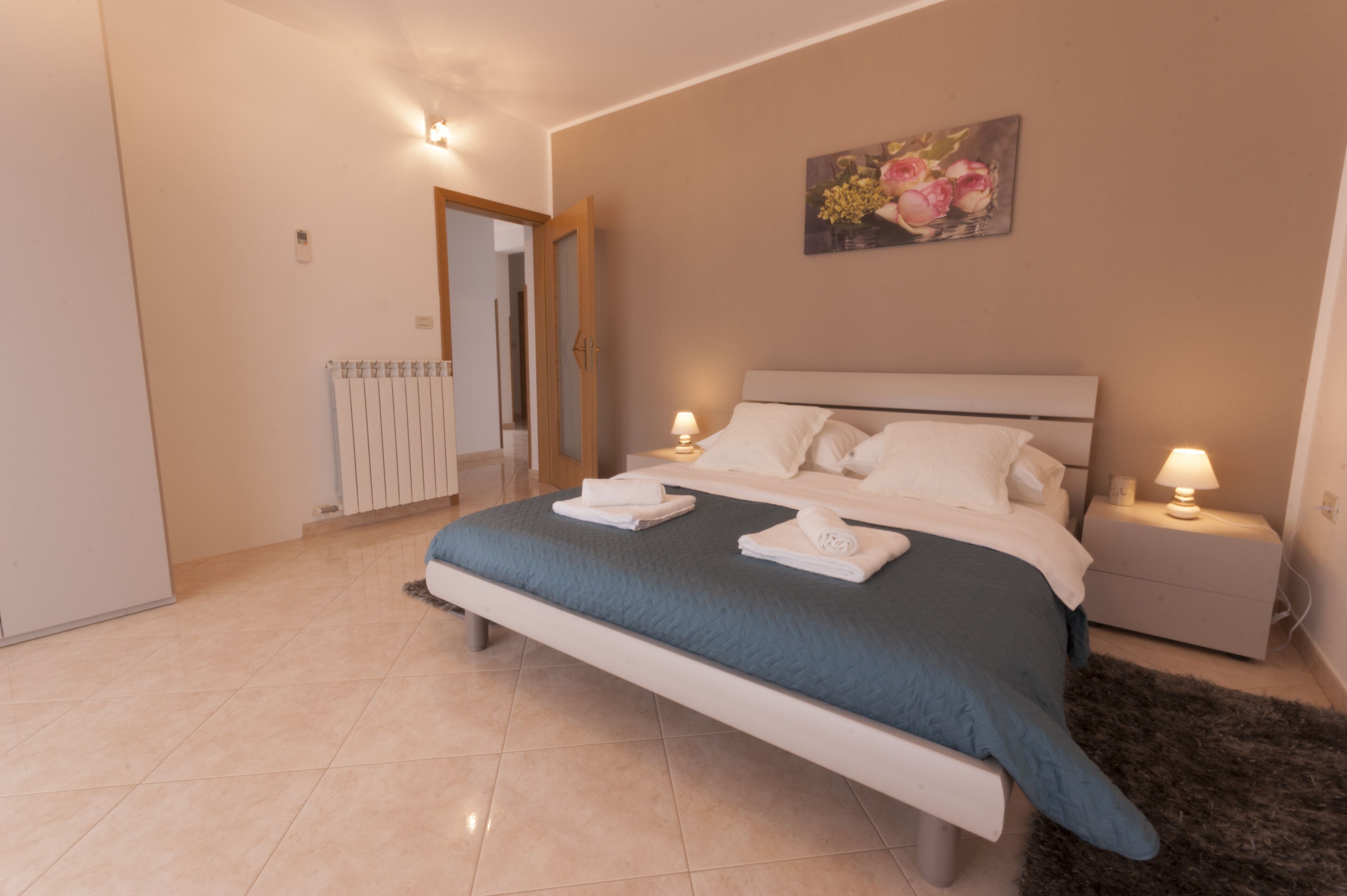 A bedroom with a bed, two bedside tables with lamps, a radiator, and a painting on the wall.