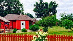 Una casa roja con puerta blanca, rodeada por una valla de piquetes rojos, un césped y varios árboles.