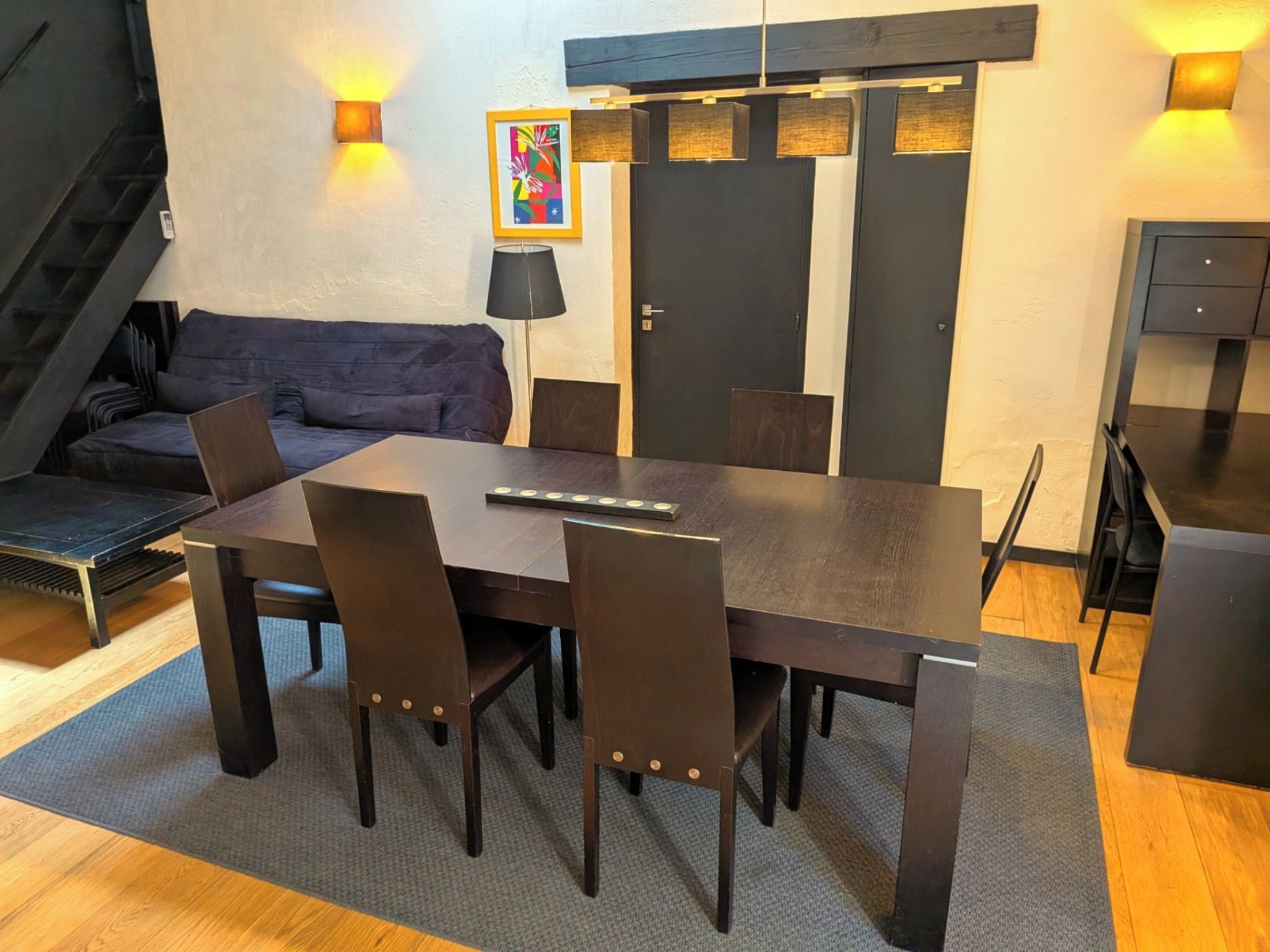 A dining area with a rectangular table, four chairs, a sofa, and a staircase.