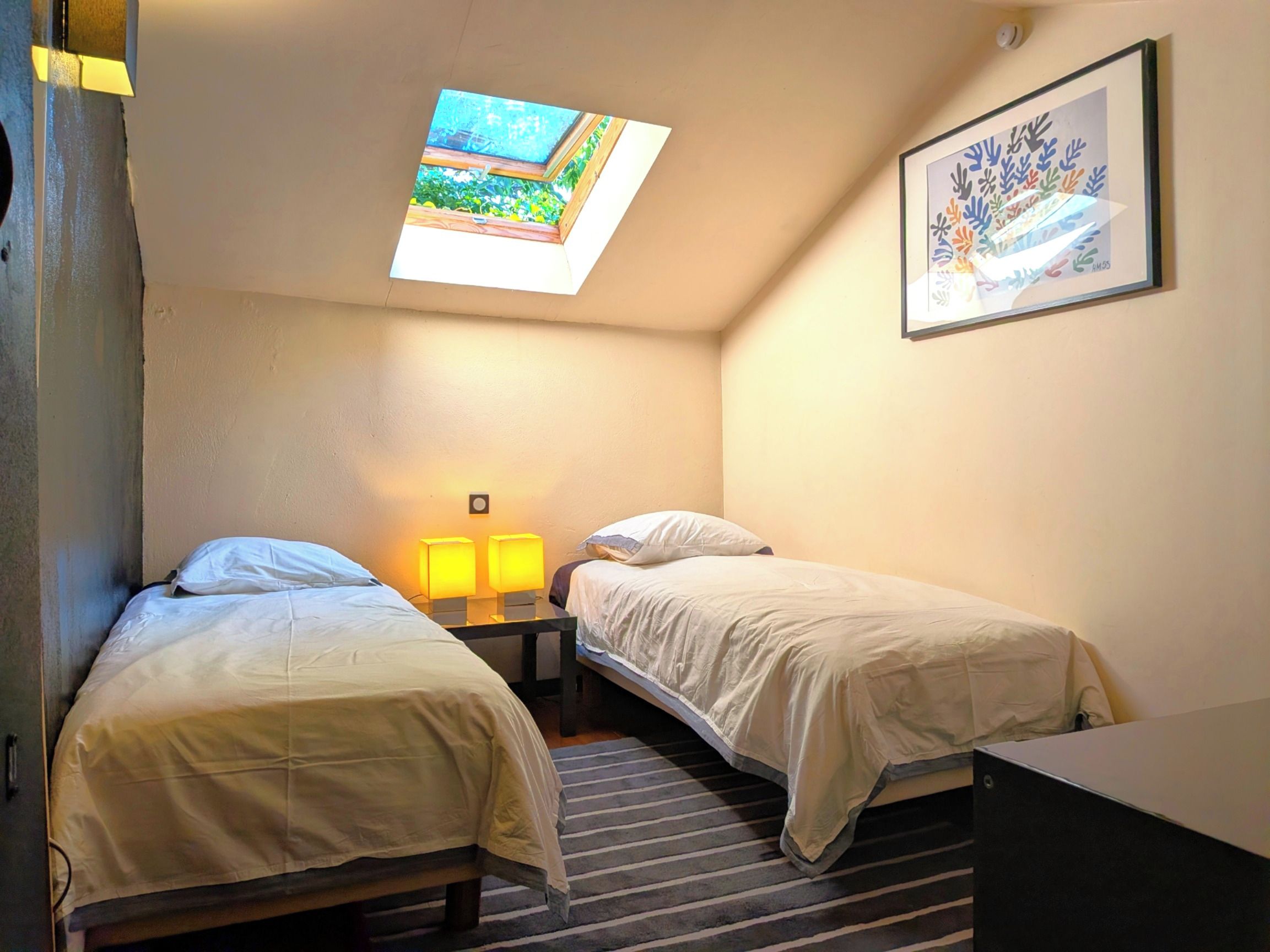 A small, cozy room with two single beds, a nightstand with a lamp, a framed picture on the wall, and a skylight above the beds.