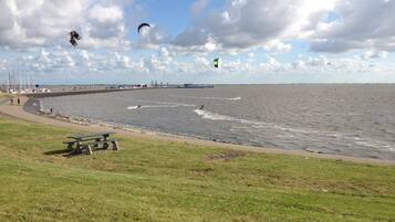 Een kustgebied met een grasveld voor, een picknicktafel en een uitzicht op zee, met mensen en vlieger(s) op de achtergrond.