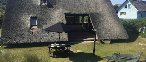 Uma casa com fachada de palha única, uma pequena janela e porta que dá para área externa com mesa de piquenique e cadeiras de descanso.