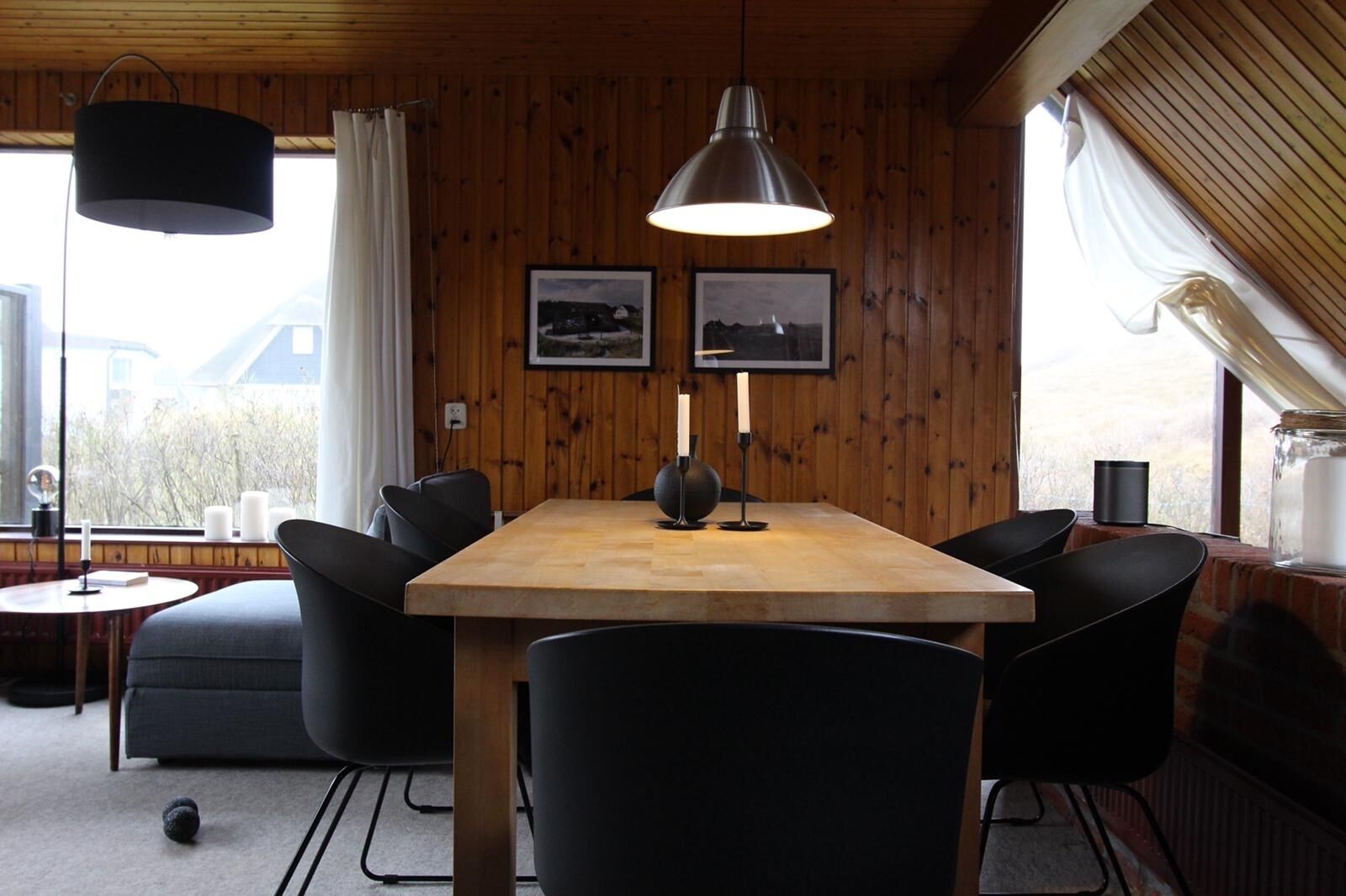 Een kamer met houten lambrisering, een houten tafel, zwarte stoelen en een hanglamp. Twee ingelijste foto's hangen aan de muur, er is een raam met gordijnen en een klein tafeltje met een kaars en een boek.