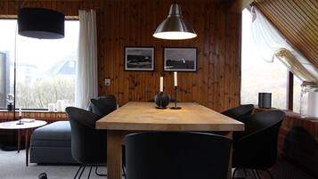 Een kamer met houten lambrisering, een houten tafel, zwarte stoelen en een hanglamp. Twee ingelijste foto's hangen aan de muur, er is een raam met gordijnen en een klein tafeltje met een kaars en een boek.