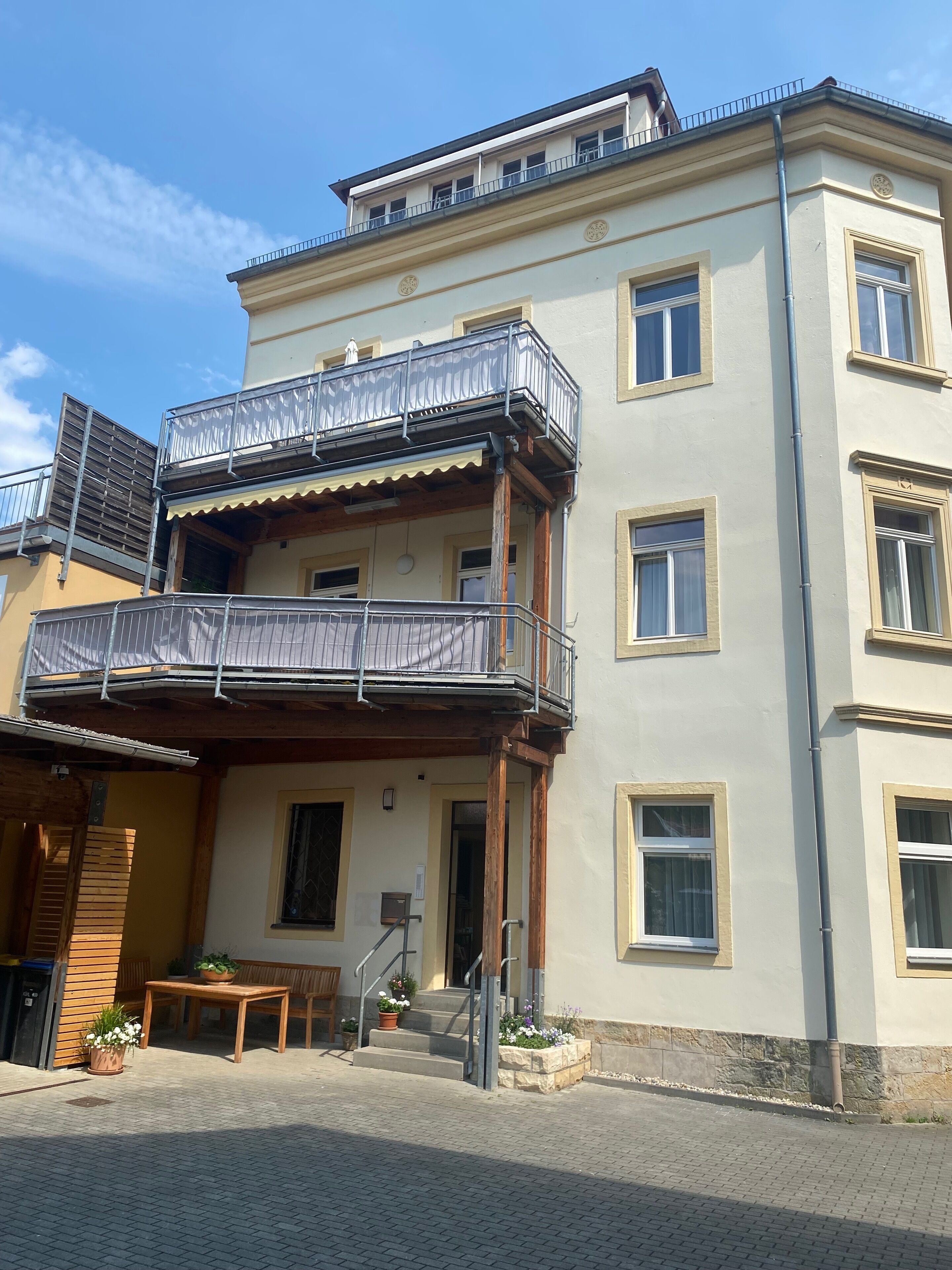 Ein mehrstöckiges Gebäude mit einem Balkon im zweiten Stock, einer Holzbank davor und einem gepflasterten Eingangsbereich.