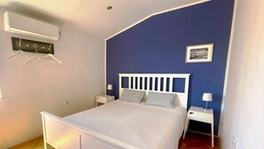 Una camera da letto con un letto bianco, due comodini, un condizionatore d'aria a parete, un pavimento in legno e una parete decorata in blu.