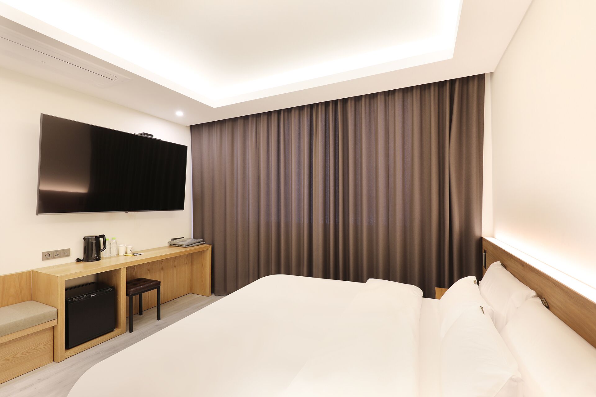 A hotel room with a bed, a TV mounted on the wall, a desk with a coffee maker, and a small refrigerator.