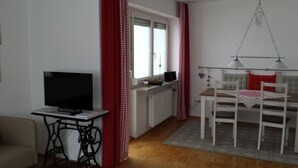 A living room with a television, a table with chairs, a window with curtains, and a radiator. - Cozy city apartment with carport for 2 to 5 people near the thermal baths (Bad Reichenhall)