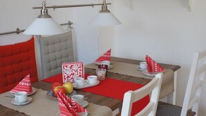 A dining area with a wooden table, white chairs, and a red table runner. There are two hanging lamps, a shelf with mugs and books, and a red cushion on the chair. - Cozy city apartment with carport for 2 to 5 people near the thermal baths (Bad Reichenhall)
