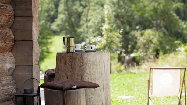 A rustic outdoor seating area with a wooden table, a metal coffee pot, and a cushion.