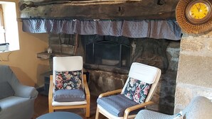 A cozy room with a stone fireplace, two wooden chairs with floral cushions, a small table, and a clock on the wall.