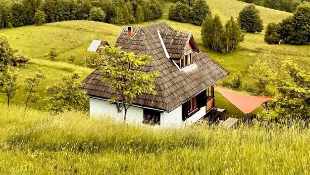 Ett hus på landsbygden med ett sluttande tak, omgivet av frodiga, gröna fält och en skogbeklädd kulle i bakgrunden.