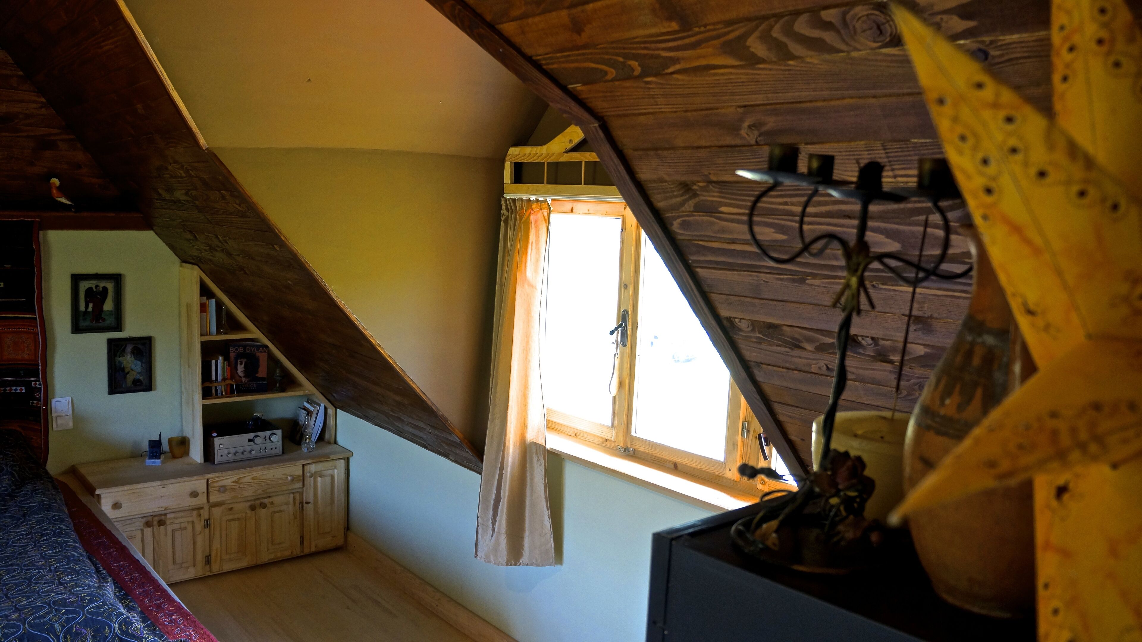Une pièce avec un plafond en pente, des poutres en bois, une fenêtre avec un rideau, un lit recouvert d’une courtepointe à motifs et une petite armoire en bois contenant divers objets.